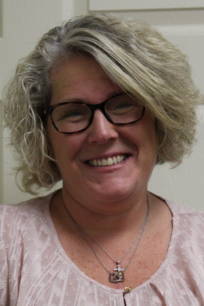 Headshot of amy lynne black, a smiling woman with wavy blonde hair, glasses, and a heart pendant necklace.