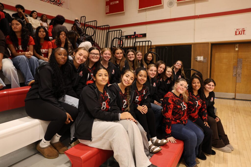 Members of the Liberty High School girls' volleyball team in black, red, and white uniforms pose together while seated on red bleachers inside the school gymnasium.